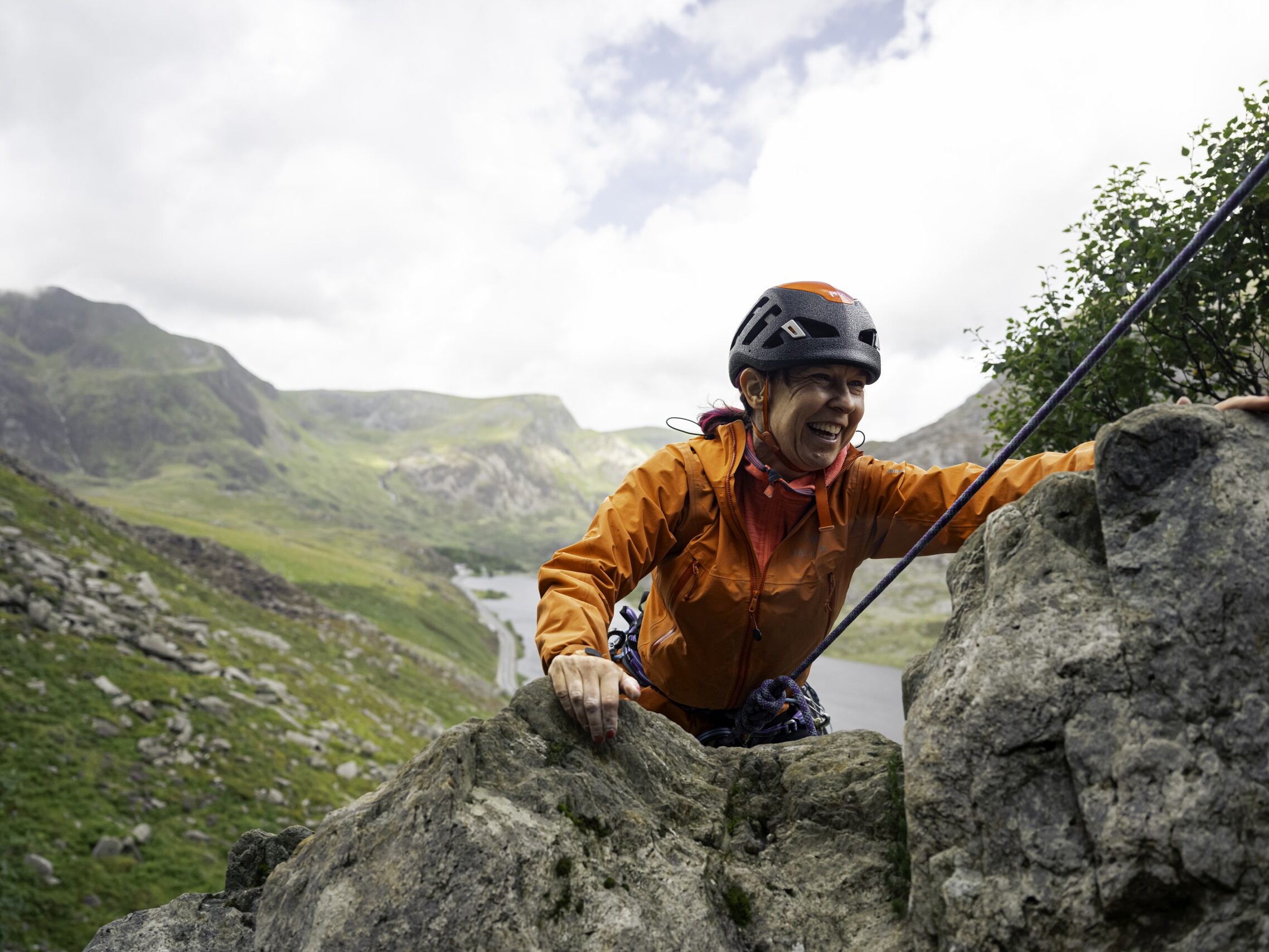 Woman with pink hair, an orange coat, and climbing helmet is seen cresting over the the top of a cliff with green hills and a lake behind her.