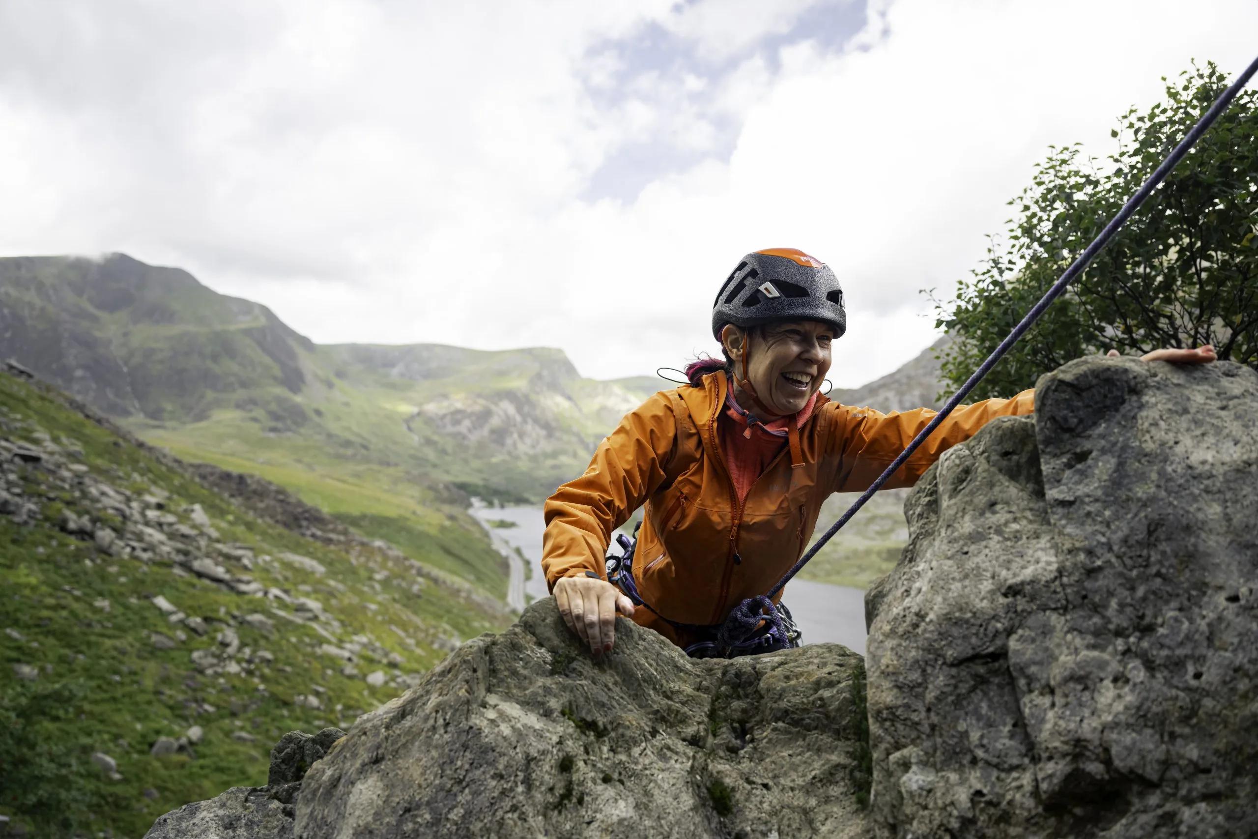 Woman with pink hair, an orange coat, and climbing helmet is seen cresting over the the top of a cliff with green hills and a lake behind her.